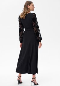 Schwarzes Kleid mit langen, durchsichtigen Spitze-Ärmeln, die mit floralen Mustern verziert sind. Das Kleid hat eine taillierte Taille und fällt bis zur Midi-Länge.