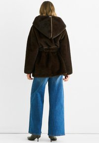 Brown faux fur jacket with a large hood, belted waist, and wide sleeves, paired with high-waisted blue jeans and green heeled boots.