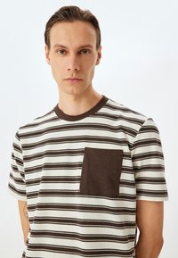 Stribet t-shirt i hvide og brune nuancer, med en brun paspel lomme. Bomuldsmateriale med rund halsudskæring og korte ærmer.