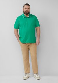 Polo vert avec des bordures blanches, associé à un pantalon beige et des baskets blanches. La chemise présente un col classique et un logo discret.