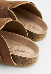 Brune slip-on sandaler med en korksål og en semilær tekstur. Har åbne sider og en syet rem med en spænde. Rød branding indeni.