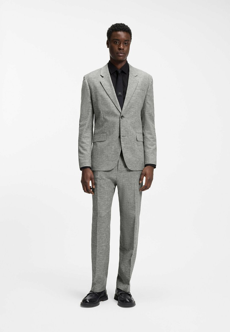 Homme debout portant un costume gris avec une chemise noire et des chaussures noires sur un fond blanc uni.