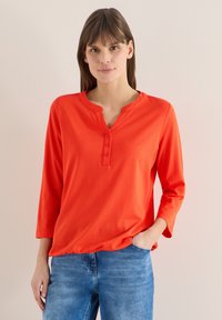 Femme portant un haut henley à manches longues de couleur orange vif avec trois boutons, associée à un jean en denim bleu, debout devant un fond neutre.