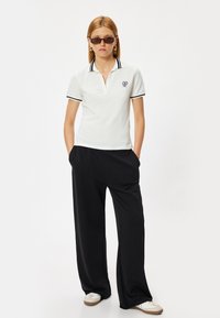 Witte polo met zwarte kraag en mouwstrepen, voorzien van een klein logo. Gecombineerd met losse zwarte broeken en witte sneakers.