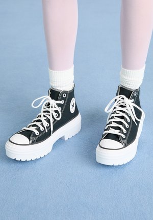 Sorte høj-top sneakers med hvide såler og snørebånd, med et cirkulært logo på siden og kontrasterende sorte syninger. Tekstureret overdækning.