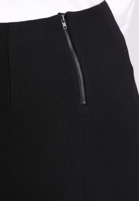 Nahaufnahme eines schwarzen, strukturierten Stoffrocks mit einem offenen silbernen Reißverschluss an der Seite, getragen mit einem weißen Oberteil.