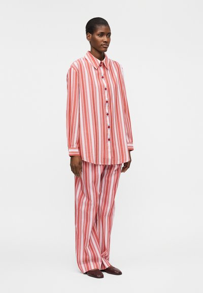 Naine, kes kannab lahtist punase ja valge vertikaalsete triipudega särki ning sobivaid laiade säärtega pükse, seisab käed külgedel.