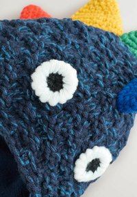 Gehaakte blauwe muts met ogen van witte en zwarte garen, met kleurrijke spikes in rood, groen en geel bovenop. Zachte textuur.
