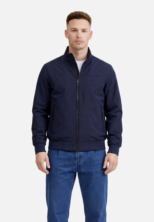 Modello maschile che indossa una giacca blu navy con zip e tasche laterali, e jeans blu, in piedi contro uno sfondo chiaro semplice.