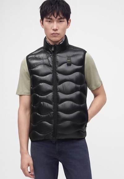 Hombre con un chaleco acolchado sin mangas negro sobre una camiseta de manga corta verde claro y vaqueros azul oscuro, de pie frente a un fondo liso.