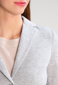 Lichtgrijze jas met een kraag, gemaakt van zachte stof, gelaagd over een lichtbeige top. De stof lijkt glad met een subtiele textuur.