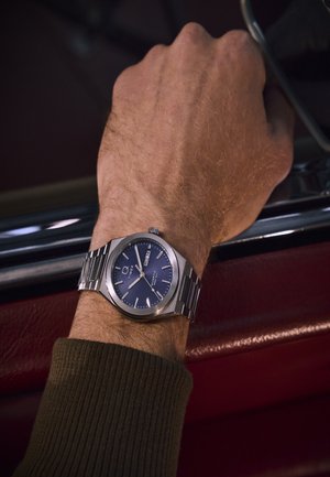 Vyro riešas su sidabriniu metaliniu laikrodžiu, rodančiu laiką ir dieną, ilsisi ant raudonos odinės automobilio durų panelės.