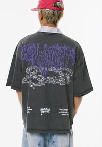 Overgroßes schwarzes T-Shirt mit kurzen Ärmeln, das lila grafischen Text und Ketten zeigt, mit ausgefransten Kanten und zusätzlichem Text, der darunter gedruckt ist.