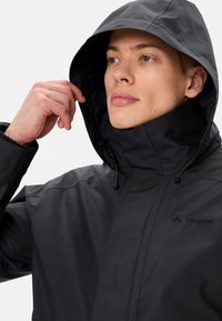 Giacca nera impermeabile con colletto alto e cappuccio regolabile. Caratterizzata da un tessuto liscio, cuciture minime e un logo discreto sul petto.
