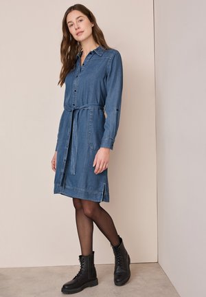 Abito camicia in denim di blu medio, con bottone frontale, vita regolabile con cintura, maniche lunghe e tasche laterali. Abbinato a stivali neri con lacci.