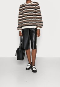 Modell iförd en randig brun och svart tröja med ballongsärmar, tillsammans med svarta lädershorts och loafers, som håller en svart väska.