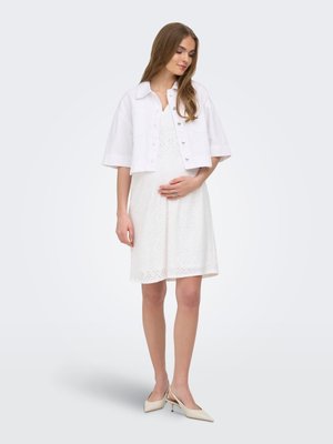 Jeune femme en robe blanche brodée anglaise, veste blanche courte à boutons et escarpins slingback beiges, tenant doucement son ventre, debout sur un fond uni.
