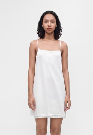 Jeune femme à la peau moyenne et aux cheveux bouclés foncés, portant une robe nuisette blanche sans manches, debout devant un fond clair uni.