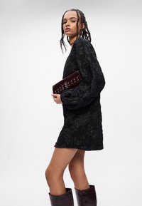 Melna izšūta mini kleita ar teksturētu ziedu rakstu, garām puff piedurknēm, savienota ar bordo taisnstūra somiņu un tumšām ceļgalu augstām zābakiem.