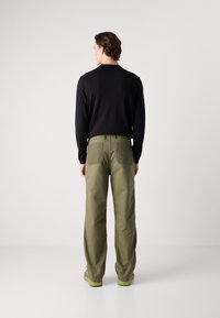 Camisa negra de manga larga, pantalones anchos color verde oliva con dos bolsillos traseros, usados con zapatos grises y amarillos, sobre un fondo liso.