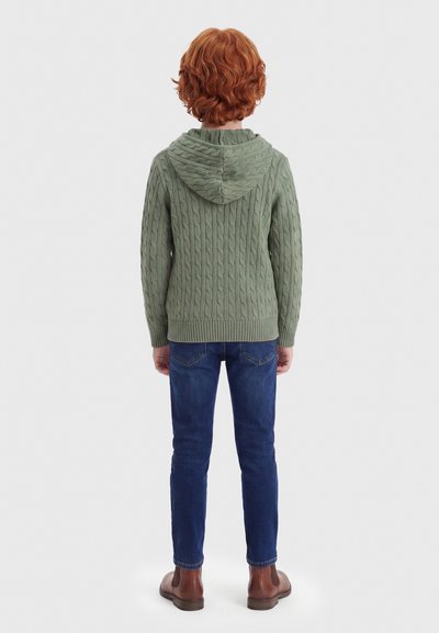 Παιδί με κόκκινα σγουρά μαλλιά που φοράει πράσινο πλεκτό πουλόβερ με κουκούλα, μπλε τζιν και καφέ μπότες, στέκεται γυρισμένο με την πλάτη σε λευκό φόντο.