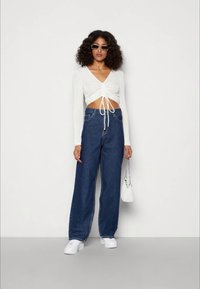Mulher com cabelo cacheado, vestindo um top cropped branco com pregas de mangas longas, jeans azuis de perna larga, ténis brancos, óculos de sol e segurando uma pequena mala branca.