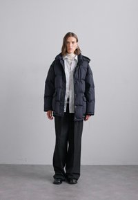 Pufferjack in donker marineblauw, voorzien van een capuchon, een voorkant rits en zijzakken. Gemaakt over een witte blouse met kraag en zwarte wijde broek.