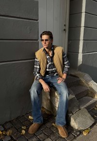 Noor inimene päikeseprillidega, ruudulise särgi, beežist tikandiga vestiga ja teksadega, istub päikesepaistelisel ajal hoone seina kõrval õues kivist treppidel.