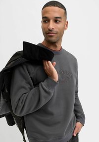 Szary sweatshirt z okrągłym dekoltem i haftowanym napisem "Originals Studio". Model trzyma czarną kurtkę z kołnierzem ze sztruksu na jednym ramieniu.