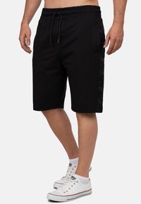 Shorts de sport noirs avec une taille élastique, cordon de serrage et poches latérales. Présente un motif de logo ton sur ton sur le côté.