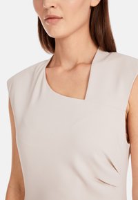 Lichtbeige mouwloze top met een ronde halslijn, een gladde textuur en drapering aan de zijkant, gemaakt van een rekbare stof.