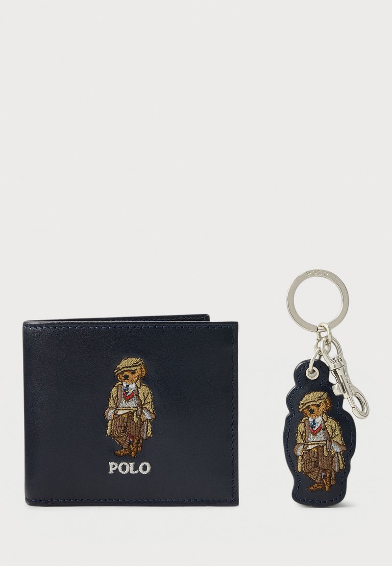 Portofel din piele bleumarin cu un mascot brodat, asociat cu un breloc din același design. Portofelul are detalii de cusătură; brelocul include un carabină.