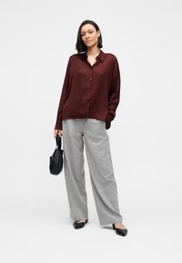 Chemise boutonnée bordeaux à coupe décontractée, associée à un pantalon large gris clair. Chaussures plates noires et petit sac à main noir complètent la tenue.