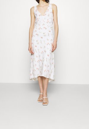 Kobieta ubrana w białą, bezrękawnikową sukienkę midi z kwiatowym nadrukiem oraz białe sandały na cienkich paseczkach na jednolitym białym tle.