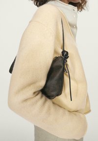 Maglione beige lavorato a maglia con vestibilità ampia, abbinato a una piccola borsa in pelle nera con chiusura a coulisse e superficie testurizzata.