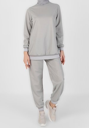 Kobieta ubrana w jasnoszary dres z paskami na mankietach i dole, beżowe sneakersy, stojąca na jednolitym jasnym tle.