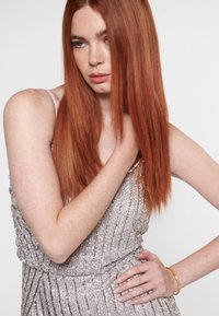 Korálkované šaty s odstíny stříbrné a růžové, s vertikálními pruhy a přiléhavým střihem. Modelka má dlouhé červené vlasy a nosí zlatý náramek.