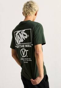 Camiseta de algodón verde oscuro con un gran gráfico blanco en la parte trasera que presenta el texto "VANS" y "OFF THE WALL". Diseño de mangas cortas y cuello redondo.