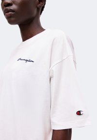 Άτομο που φοράει λευκό t-shirt Champion με κεντημένο λογότυπο στο στήθος και μικρό λογότυπο στο αριστερό μανίκι.
