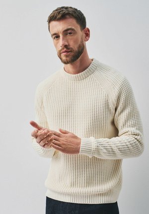 Man with short brown hair and beard wearing a cream textured knit sweater, standing and gently rubbing his hands.