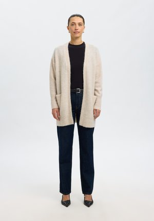 Femme debout portant un cardigan beige ouvert, un haut noir, un jean bleu foncé, une ceinture noire et des chaussures noires pointues sur un fond clair uni.