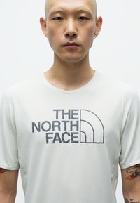 Lys grå t-skjorte med kort erm laget av teksturert stoff, med et fremtredende svart logo fra "THE NORTH FACE" på forsiden.