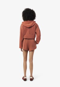 Un hoodie court de couleur rouille avec capuche et poche arrière, assorti à un short. Le tissu semble doux avec une coupe décontractée.