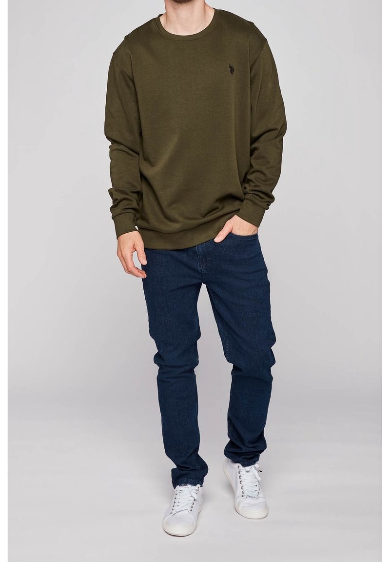 Sweat-shirt vert olive à col rond et poignets côtelés, associé à un jean bleu foncé et des baskets blanches, avec un petit logo sur la poitrine.