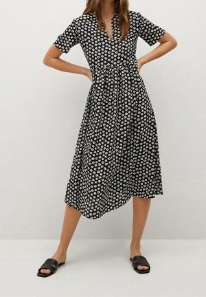 Femme portant une robe midi noire à motif floral blanc, manches courtes, col en V, et des sandales noires ouvertes à l’avant, debout les mains sur les hanches.