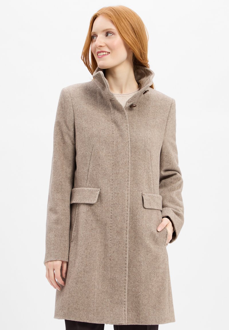 Donna con capelli rossi che indossa un cappotto di lana beige con colletto alto, bottoni frontali, tasche a patta e mani in tasca, che guarda a destra e sorride.