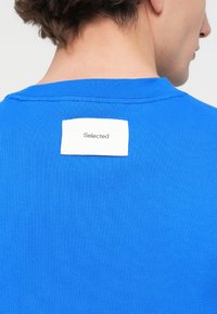 Μπλε βαμβακερό t-shirt με στρογγυλή λαιμόκοψη. Μια ετικέτα με την ένδειξη "Selected" είναι ραμμένη στην πλάτη. Λεία υφή και casual σχεδιασμός.