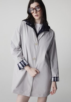 Donna che indossa un cappotto grigio chiaro con polsini e colletto a quadri blu navy, occhiali oversize con montatura tartarugata, in piedi contro uno sfondo semplice grigio chiaro.