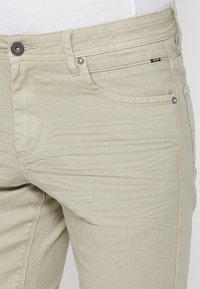 Gros plan de la taille et de la partie supérieure de la cuisse d'un jean en denim beige avec poches avant, passants de ceinture et détail du bouton en métal.