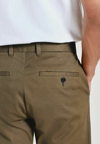 Pantalons en tissu texturé marron avec une coupe ajustée, dotés de deux poches arrière et d'un détail de bouton. Portés avec une chemise blanche.